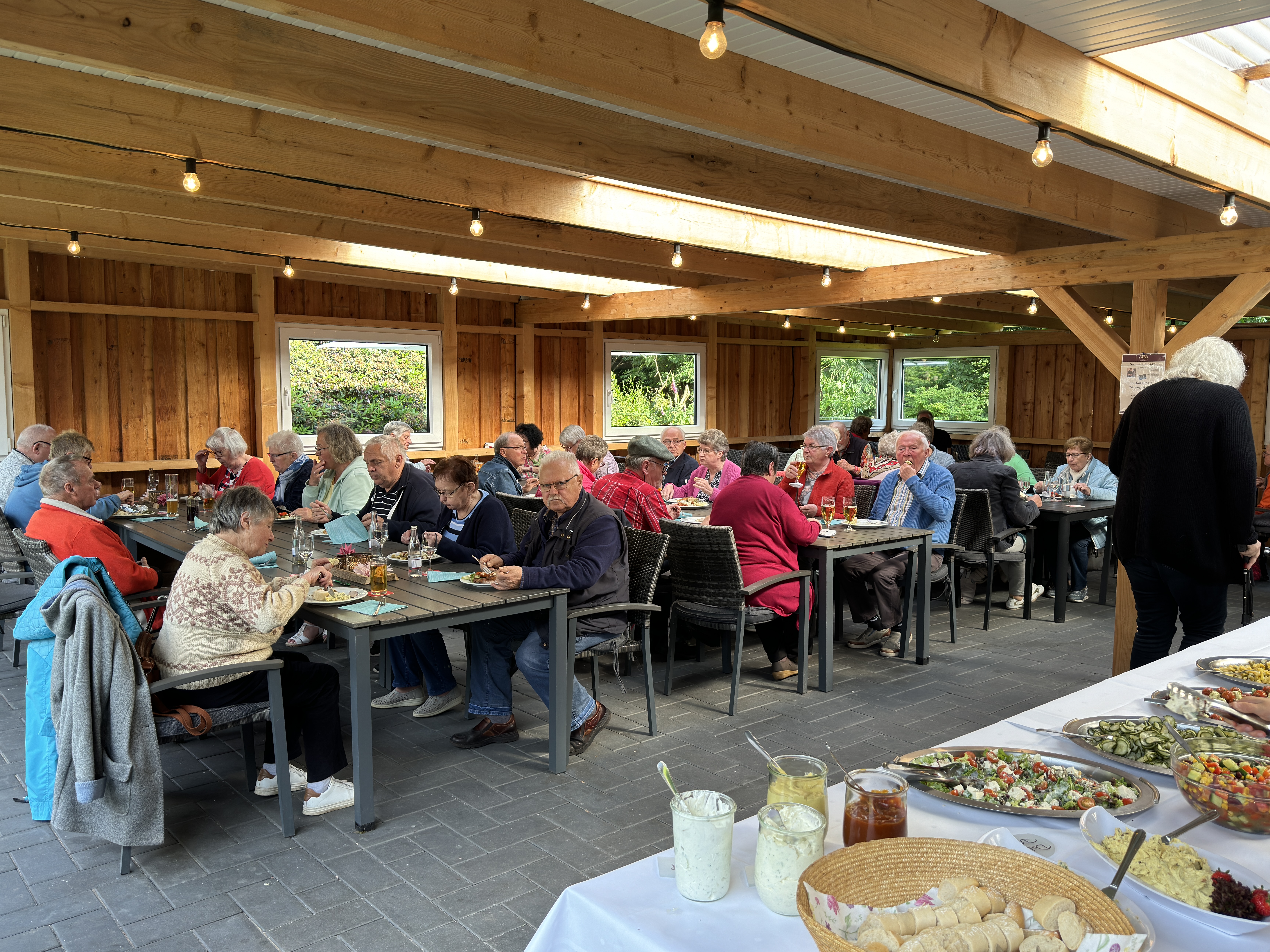 Im Vordergrund des Bildes ist ein reich gedecktes Buffet zu sehen, auf dem verschiedene Salate, Dips und Brotscheiben ansprechend angerichtet sind. Die Tische sind mit Getränken, Tellern und Besteck gedeckt, und es herrscht eine fröhliche, entspannte Stimmung.