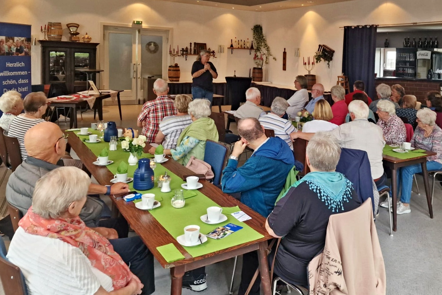 Erste Hilfe im Alter - Nach der Kaffeetafel gab es wichtige Informationen Es sind die Teilnehmerinnen und Teilnehmer des monatlichen Treffens des VdK Celle-Altstadt zu sehen. Sie sitzen an Tischen, auf denen hellgrüne Tischdecken liegen. Darauf stehen noch die Kaffeetassen. Im Hintergrund steht Christiane Gentzmann vom Deutschen Roten Kreuz.